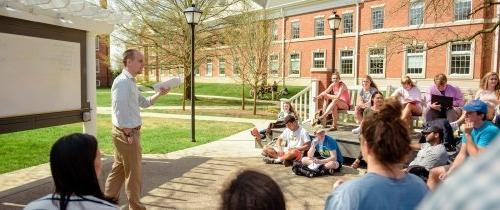 Students listening to a professor in an outdoor classroom with white board behind the professor